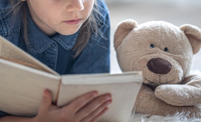 Little girl lying on the floor reads a book next to her teddy bear